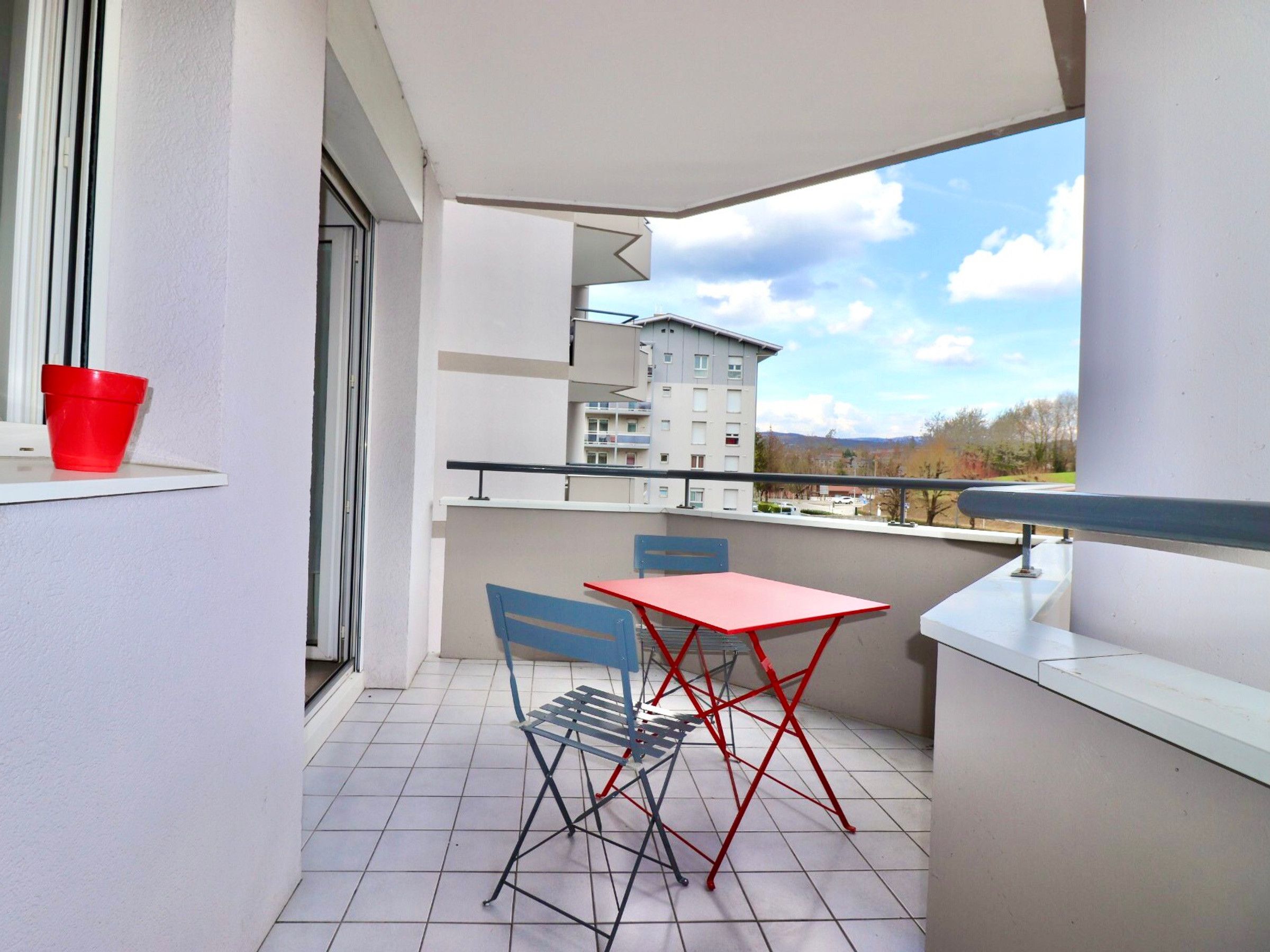 A balcony with a red table and blue chairs, a red bucket, and a view of a building and trees.