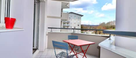 A balcony with a red table and blue chairs, a red bucket, and a view of a building and trees.