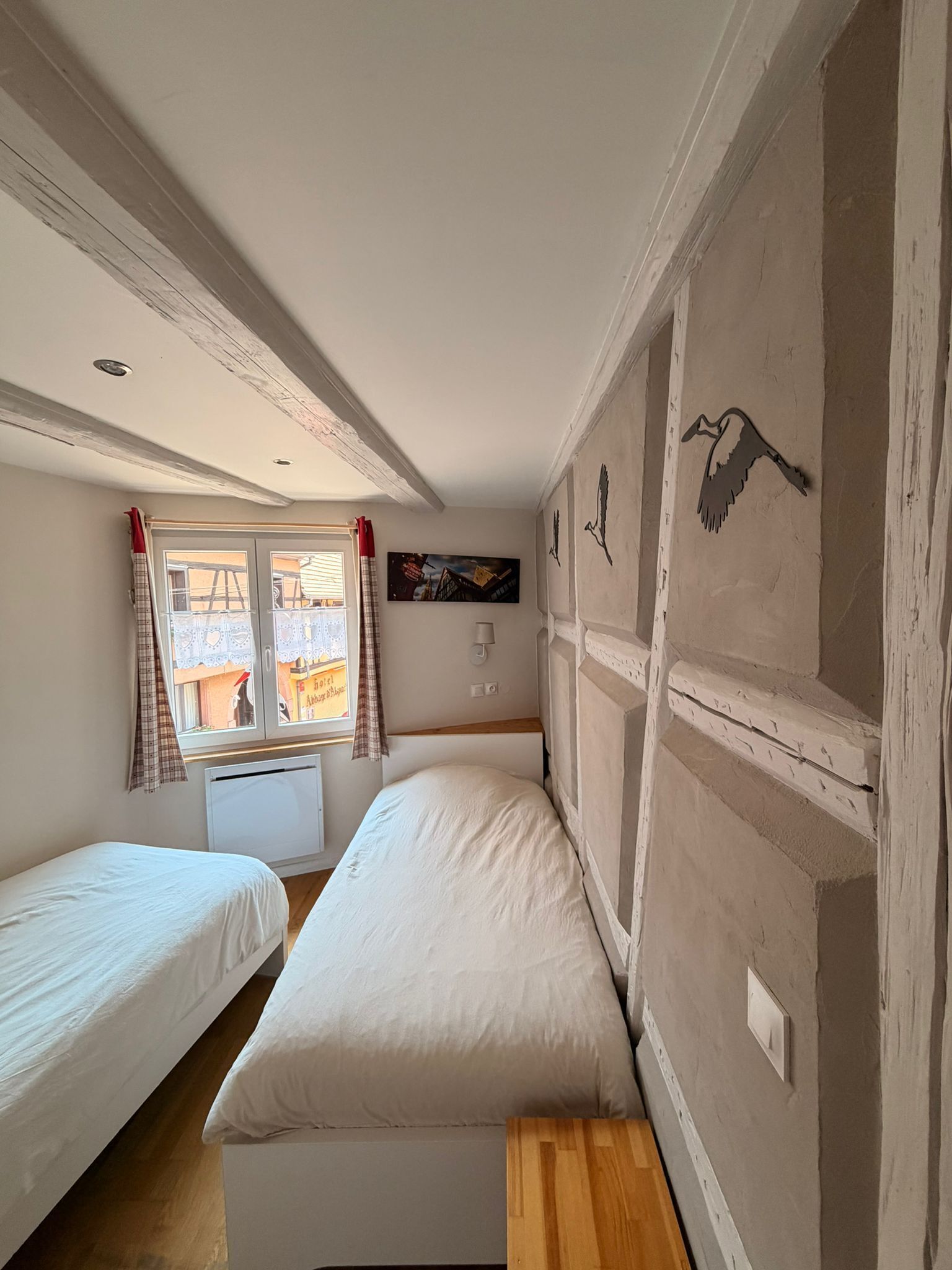 A room with two beds, a window with curtains, a wall-mounted lamp, and a painting.