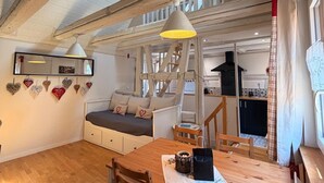 Une pièce chaleureuse de style loft, avec des poutres apparentes, un lit en mezzanine, une table à manger avec des chaises, un coin cuisine et un luminaire suspendu.