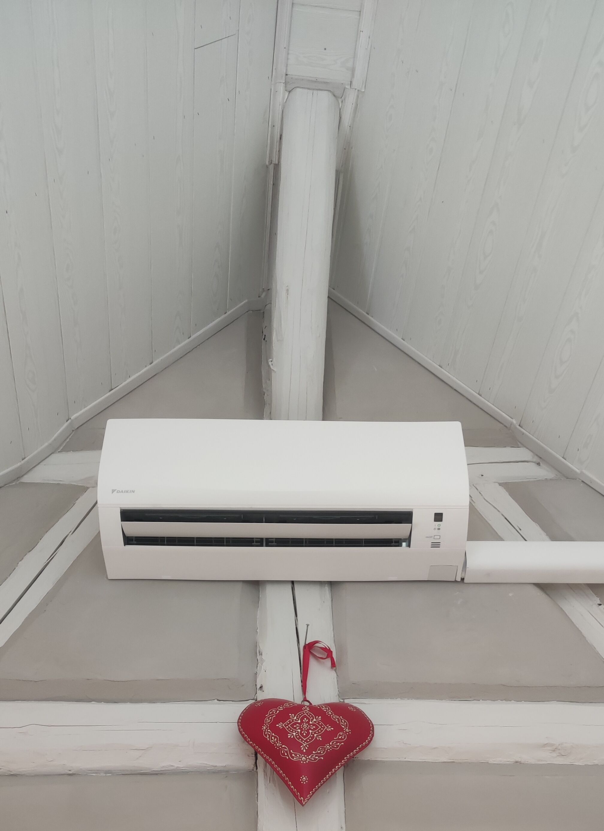 A white air conditioning unit mounted on a white wall with a red heart-shaped decoration hanging below it.