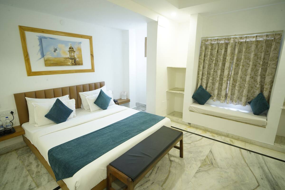 Habitación de hotel con una cama grande, un banco, una ventana con cortinas, un cuadro en la pared y una mesita con una botella.