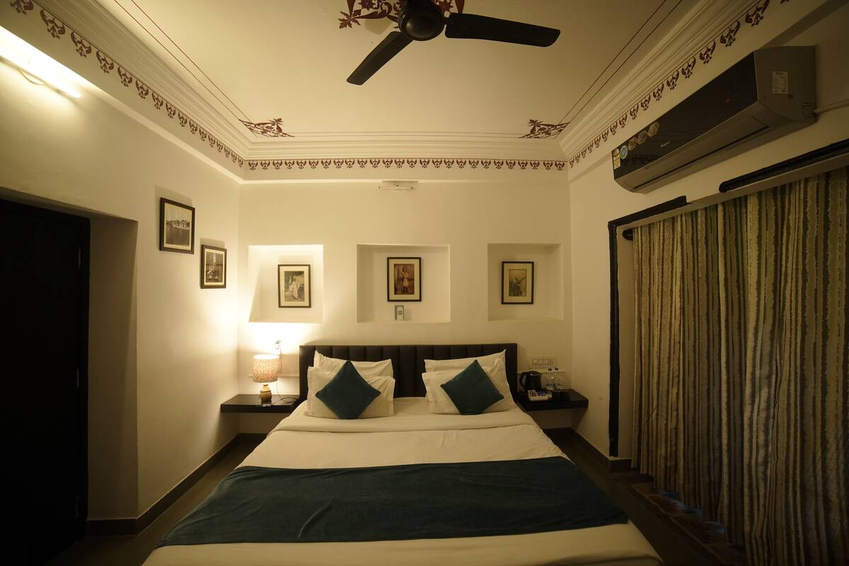 Habitación de hotel con cama, mesitas de noche, ventilador de techo, cuadros en la pared y ventana con cortinas.