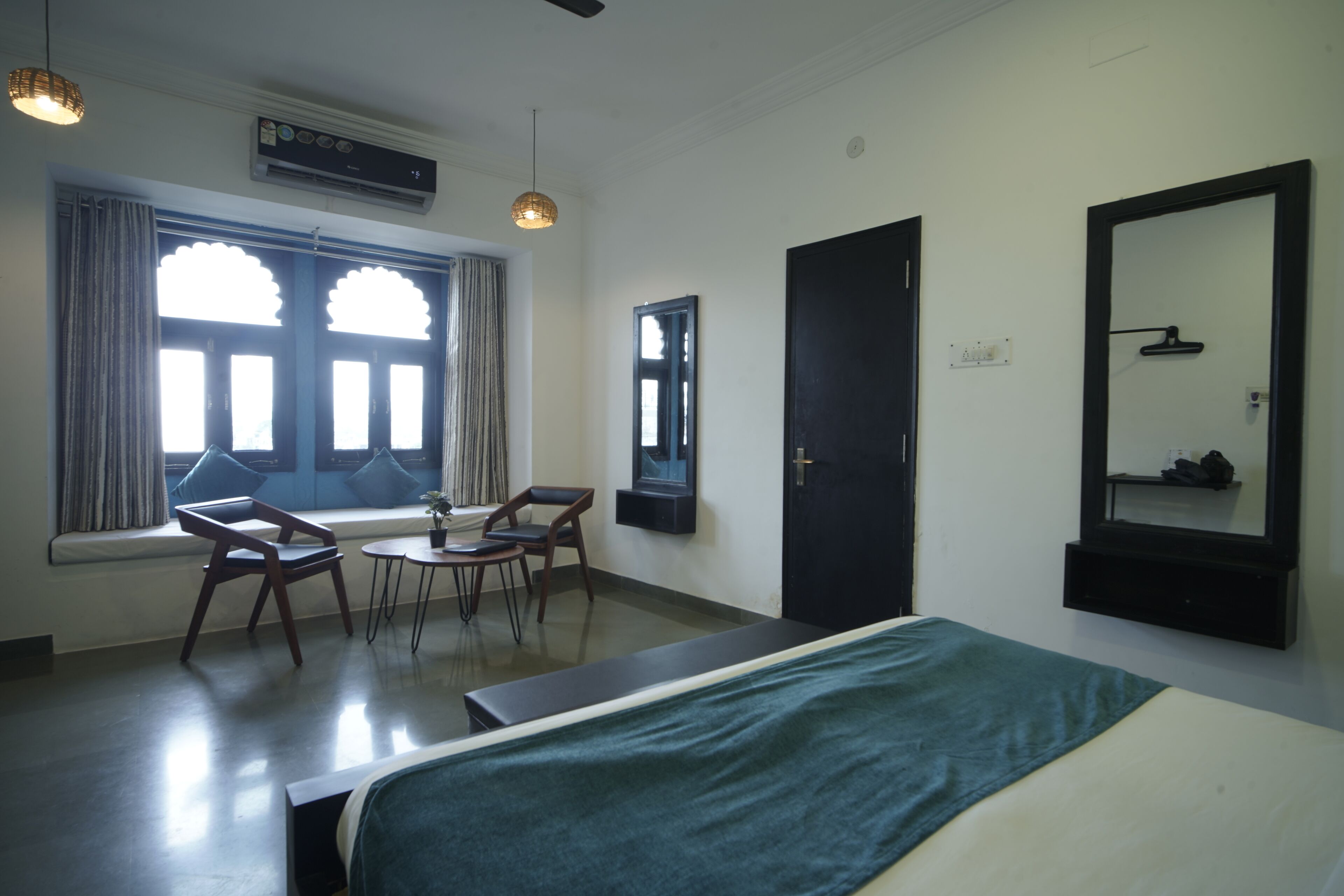 Quarto de hotel com cama, janela com cortinas, uma cadeira, uma mesinha e um espelho na parede.