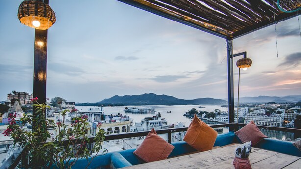 A balcony with a view of a body of water, a mountain in the distance, and a cityscape. - Banjara Heritage (Udaipur)