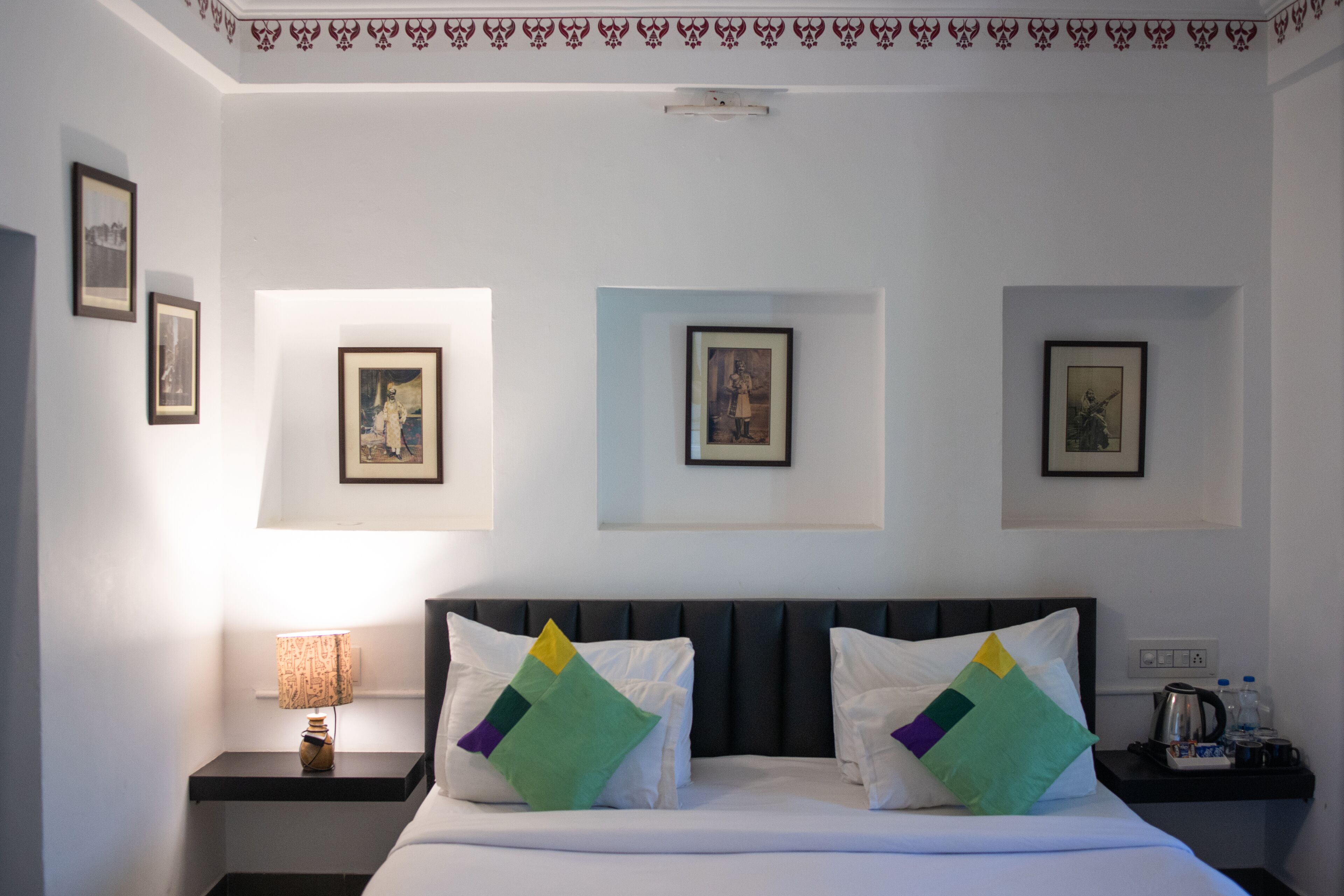 Une chambre d’hôtel comprenant un lit, deux tables de chevet, une lampe, des tableaux encadrés aux murs et un plafond orné d’un motif décoratif.
