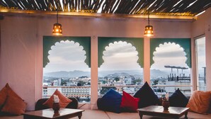 A seating area with cushioned benches and tables, a view of a cityscape through large windows, and decorative wall panels. - Banjara Heritage (Udaipur)