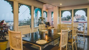 A dining area with a table set for six, surrounded by wicker chairs, with a view of a cityscape through large windows with decorative frames. - Banjara Heritage (Udaipur)