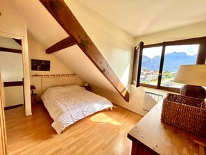 A bedroom with a sloped ceiling, a bed, a nightstand, a lamp, a window with a view of mountains, and a wicker basket on a table.