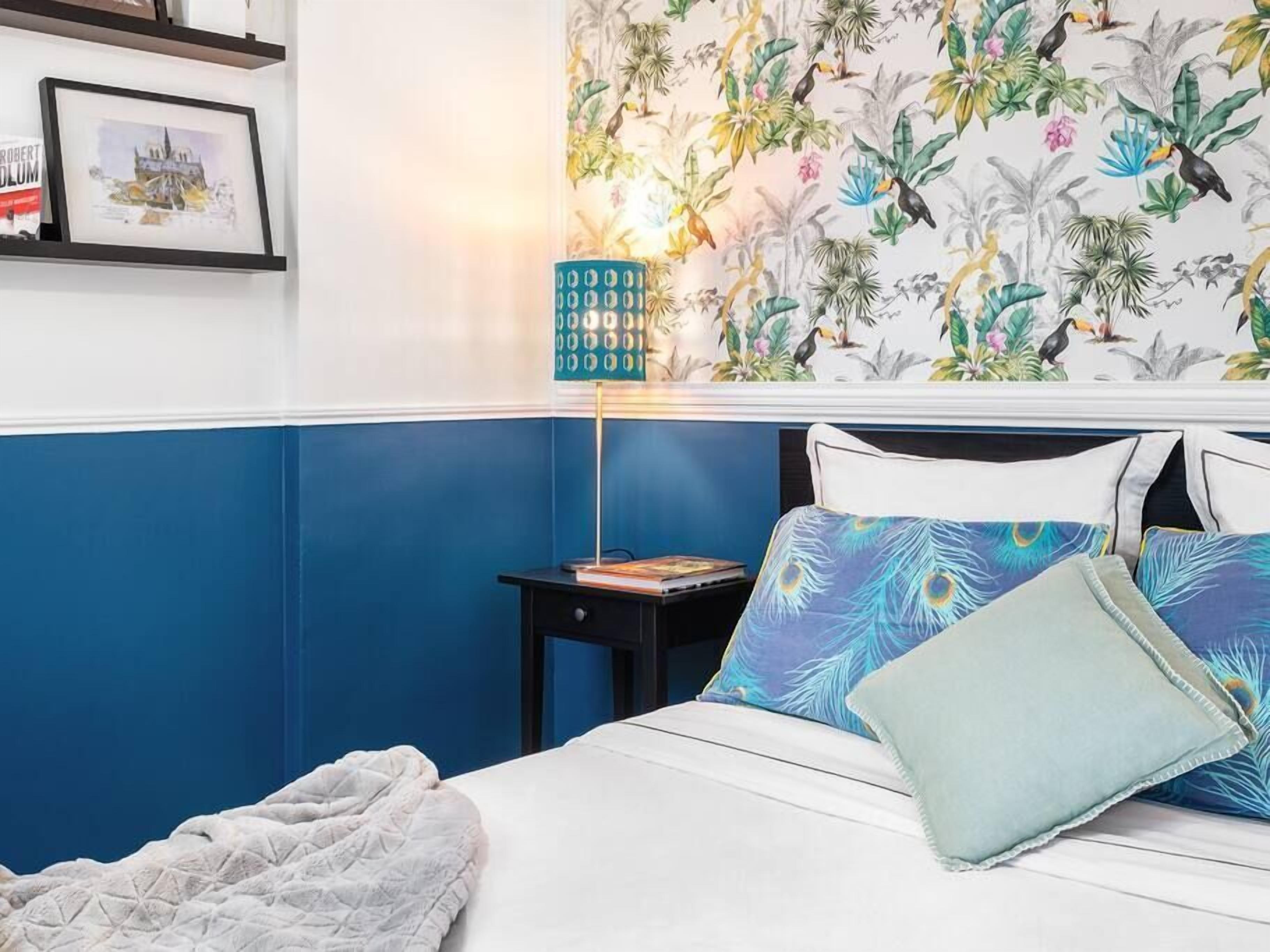 Ein Schlafzimmer mit Bett, Kissen, Nachttisch, Lampe und einer Wand mit tropischem Wandpapier.
