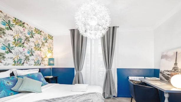 Una camera d'albergo con un letto, una scrivania, una sedia, un lampadario, tende e una parete con carta da parati tropicale.