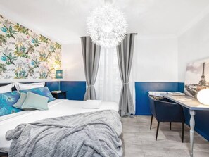 A hotel room with a bed, a desk, a chair, a chandelier, curtains, and a wall with a tropical wallpaper.