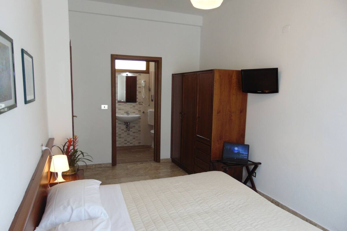 Quarto de hotel com cama, secretária com um laptop, televisão presa à parede e banheiro visível por uma porta aberta.
