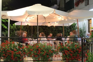 Un espacio de comedor al aire libre con sombrillas blancas y luces colgantes, rodeado de flores rojas y una barandilla de metal.