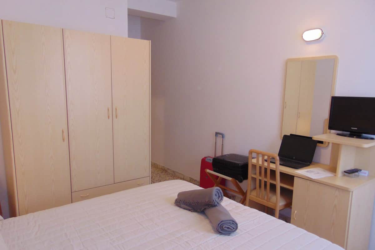 Quarto de hotel com cama, escrivaninha com um laptop, uma cadeira, mala vermelha e guarda-roupa.
