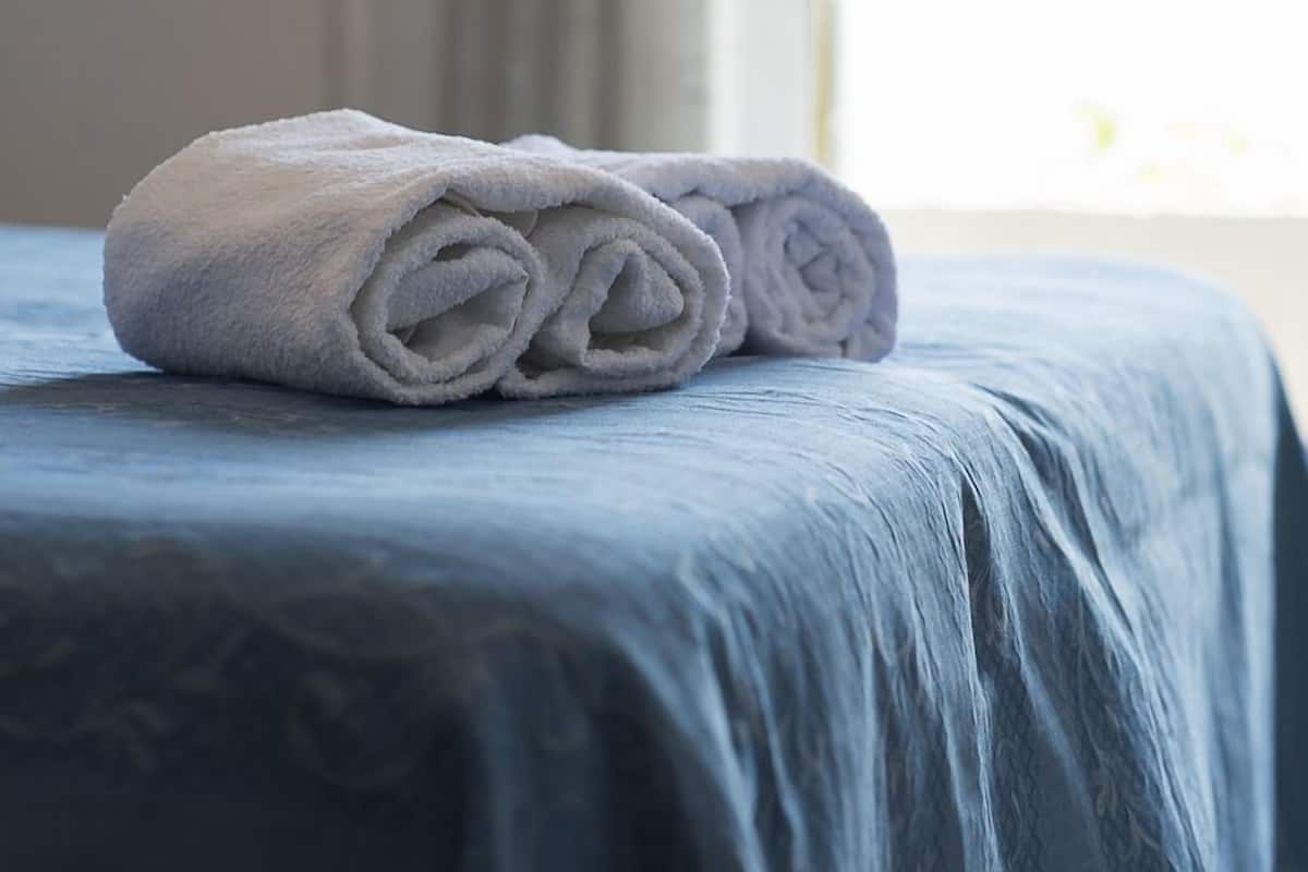 Toalha dobrada sobre a cama, com colcha azul, janela com cortinas e vista para o exterior.