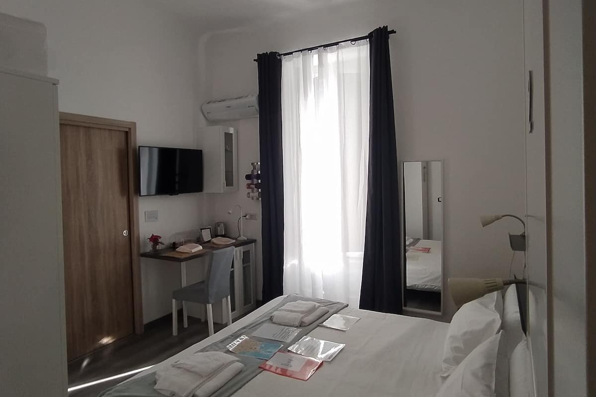 A hotel room with a bed, a desk, a chair, a TV, a mirror, and a window with curtains.