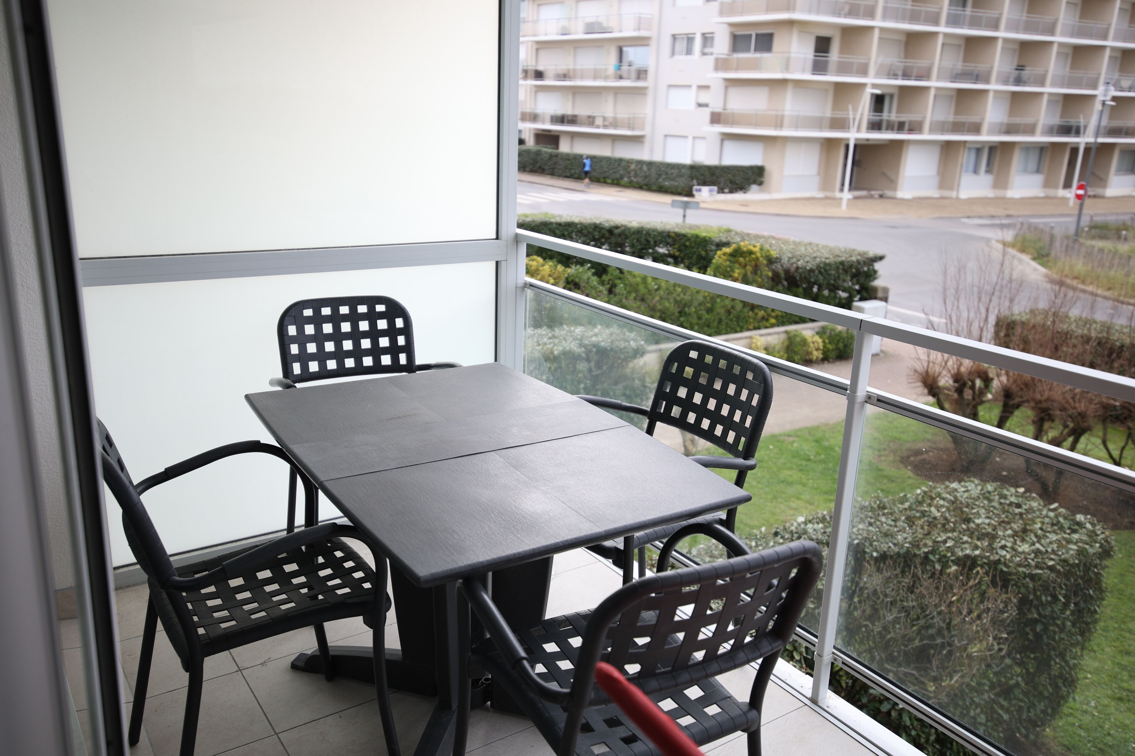 Un balcon avec une table et des chaises, donnant sur un quartier résidentiel avec une rue et des espaces verts.