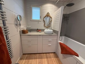 A bathroom with a wooden floor, a white vanity with a round mirror, a white sink, and a shower.