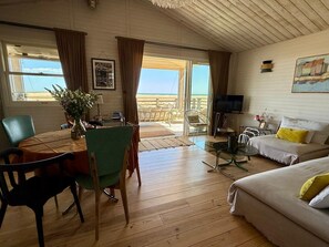 A room with a wooden floor, a dining table with chairs, a sofa, a TV, and a view of the beach.
