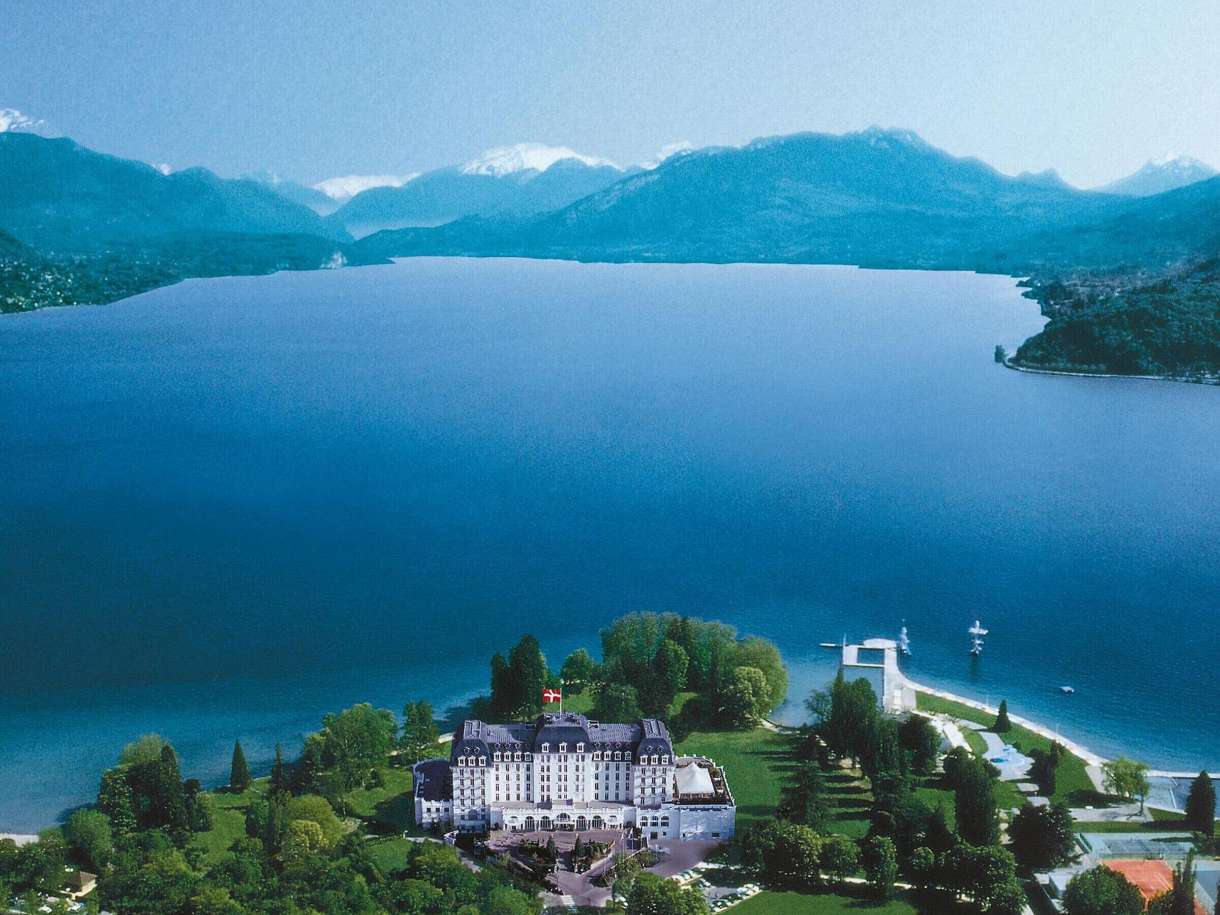 Um edifício grande, branco e de vários andares com fachada simétrica, localizado às margens de um lago com vista clara para as montanhas ao redor.