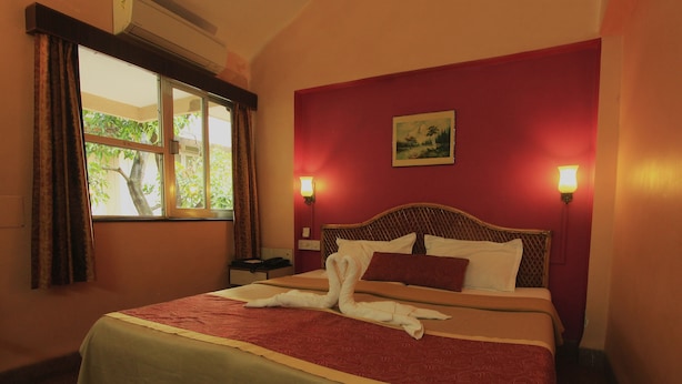 A hotel room with a bed, a nightstand, a window with curtains, and a painting on the wall. - Hotel Gopika International (Panaji)