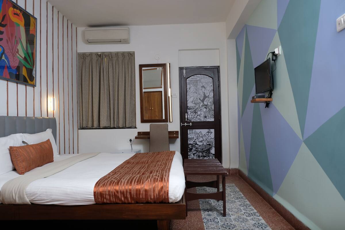 A hotel room with a bed, a desk, a chair, a TV mounted on the wall, and a door with a decorative panel.
