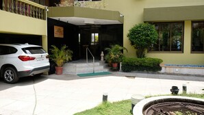 A white car parked in a courtyard with a building entrance, potted plants, and a fire pit. - Hotel Gopika International (Panaji)