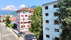 A multi-story building with balconies, a street with parked cars, and a clear view of mountains in the background. - Annecy: Spacious F2 50m from lake with balcony and parking (Annecy)