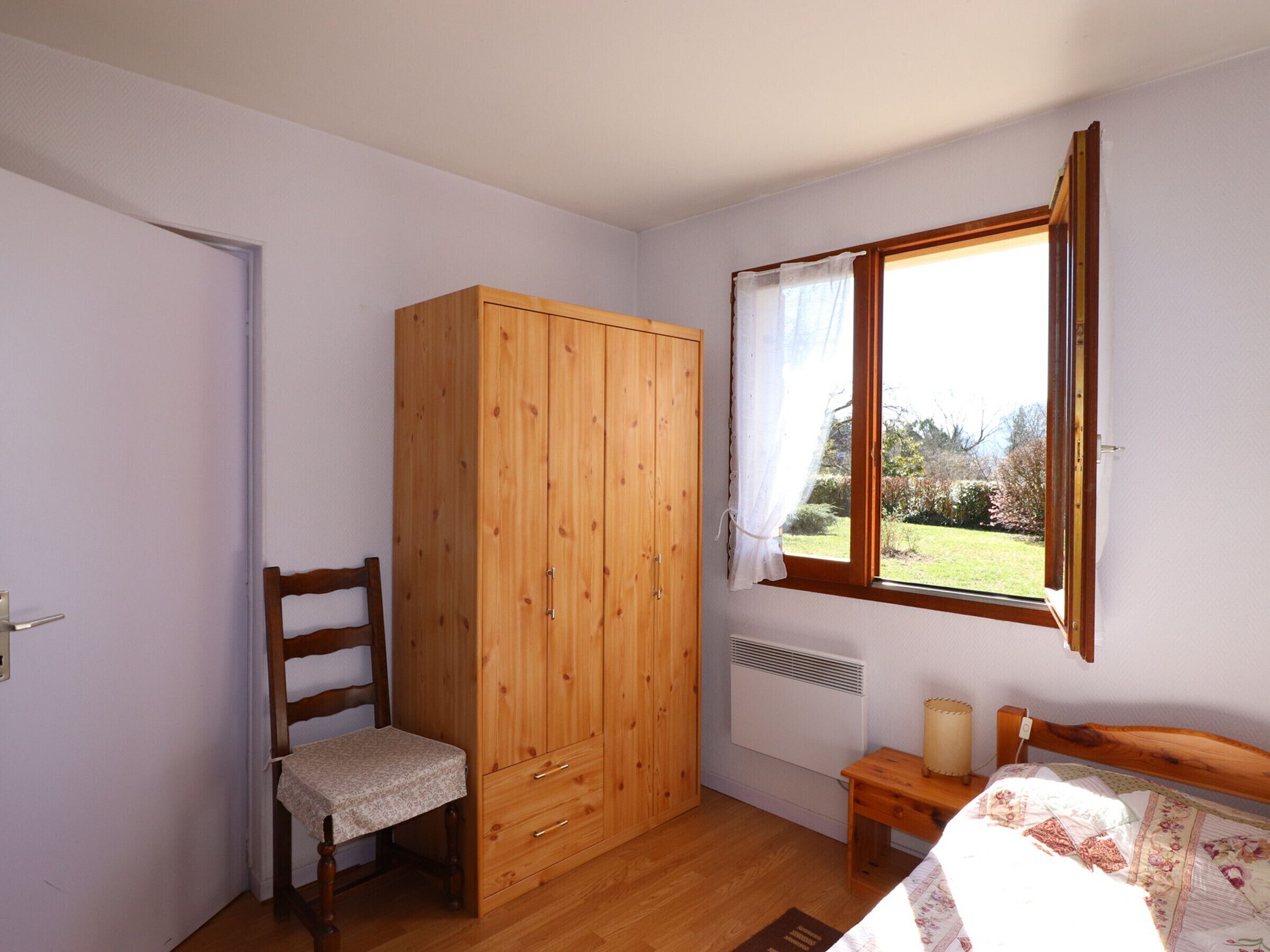 A bedroom with a wooden wardrobe, a chair, a bedside table with a lamp, and a window with a view of greenery.