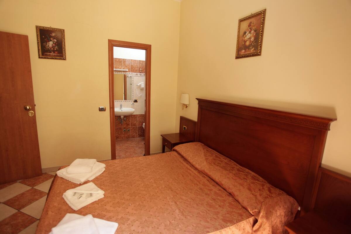 A hotel room with a bed, a wooden headboard, a nightstand, a mirror, and a door leading to a bathroom.