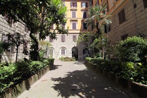 A narrow cobblestone pathway flanked by lush greenery and historic buildings with arched windows and balconies. - Elegance (Rome)