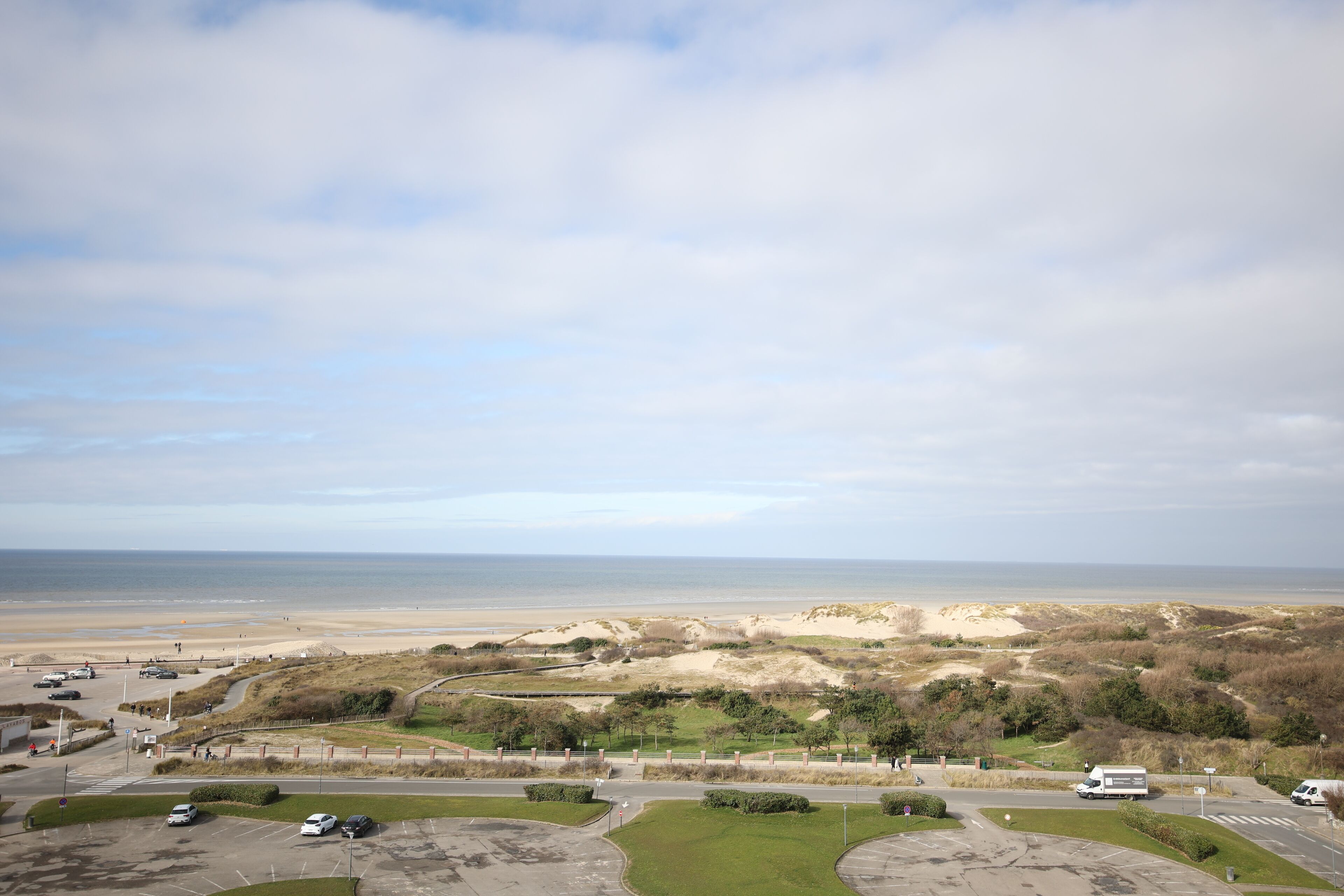 A coastal landscape with a parking area, a road, and a beach in the distance.