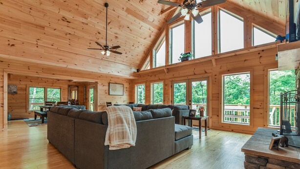 Eine geräumige Holzhütte mit einer großen, grauen Couch, einem Essbereich und zahlreichen Fenstern mit Blick auf den umliegenden Wald.