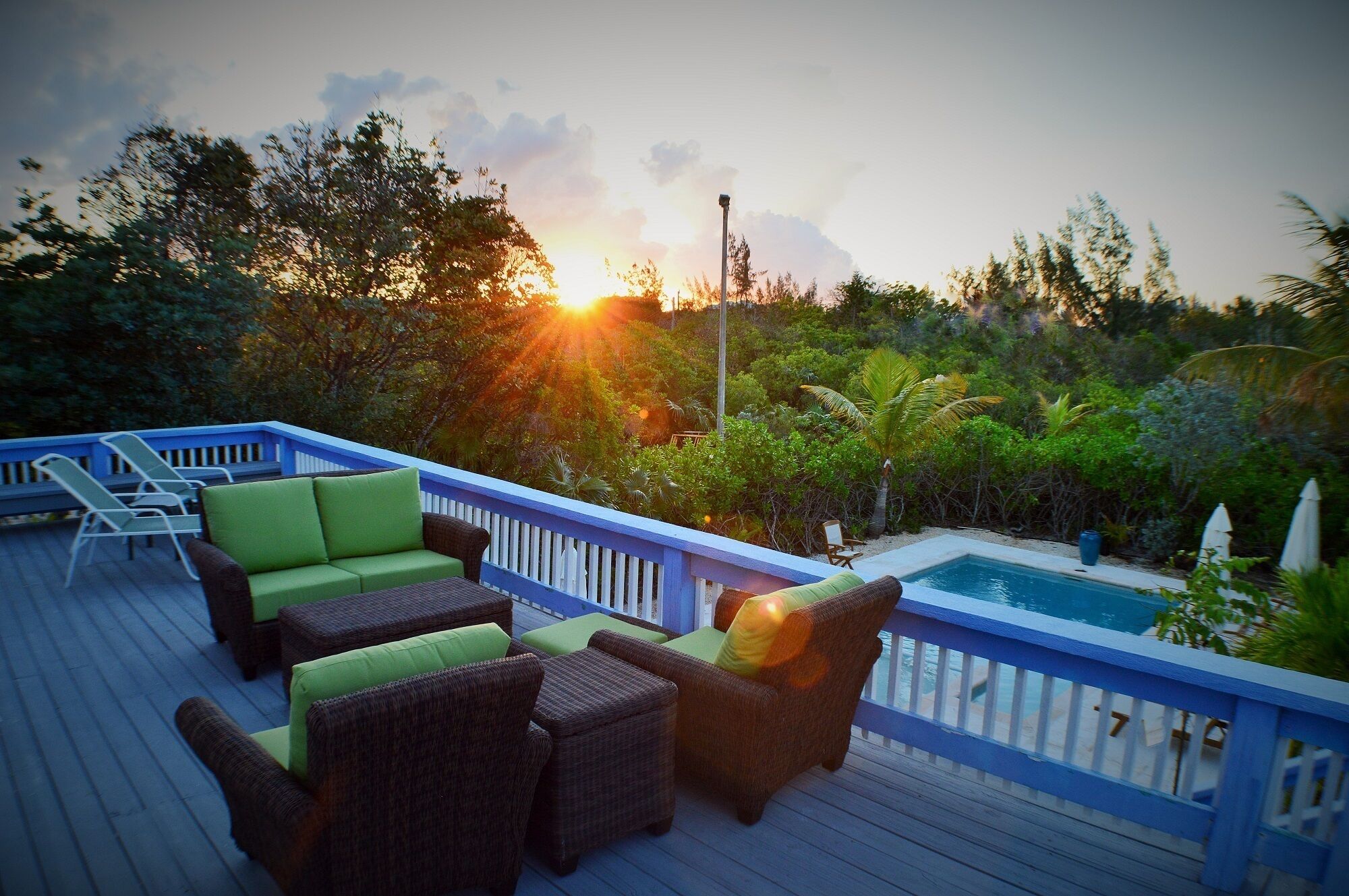 En træterrasse med fletningsmøbler og en pool, omgivet af frodig vegetation og en solnedgang.