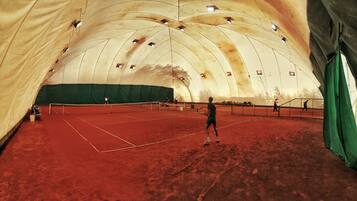 Una cancha de tenis interior con superficie de tierra batida, rodeada por una gran estructura tipo tienda con cortinas verdes.