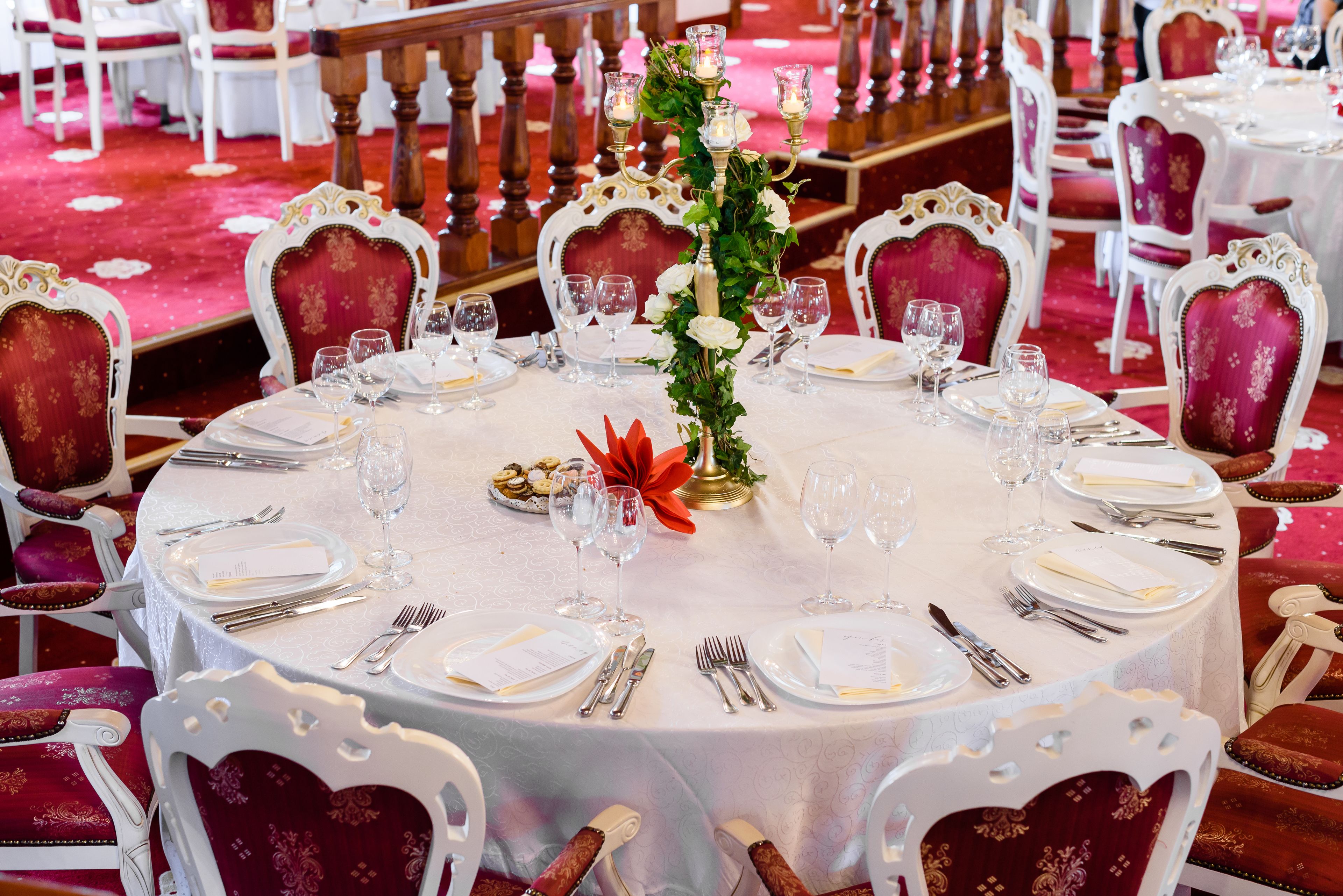 A round dining table with white tablecloth, surrounded by ornate red chairs.