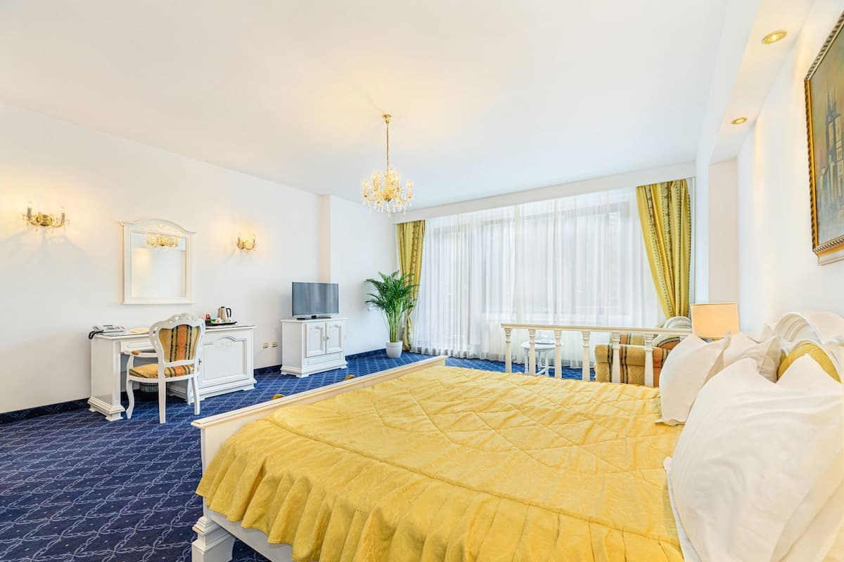 Habitación de hotel con una cama grande, un escritorio con silla, televisión, planta en maceta y ventana con cortinas.
