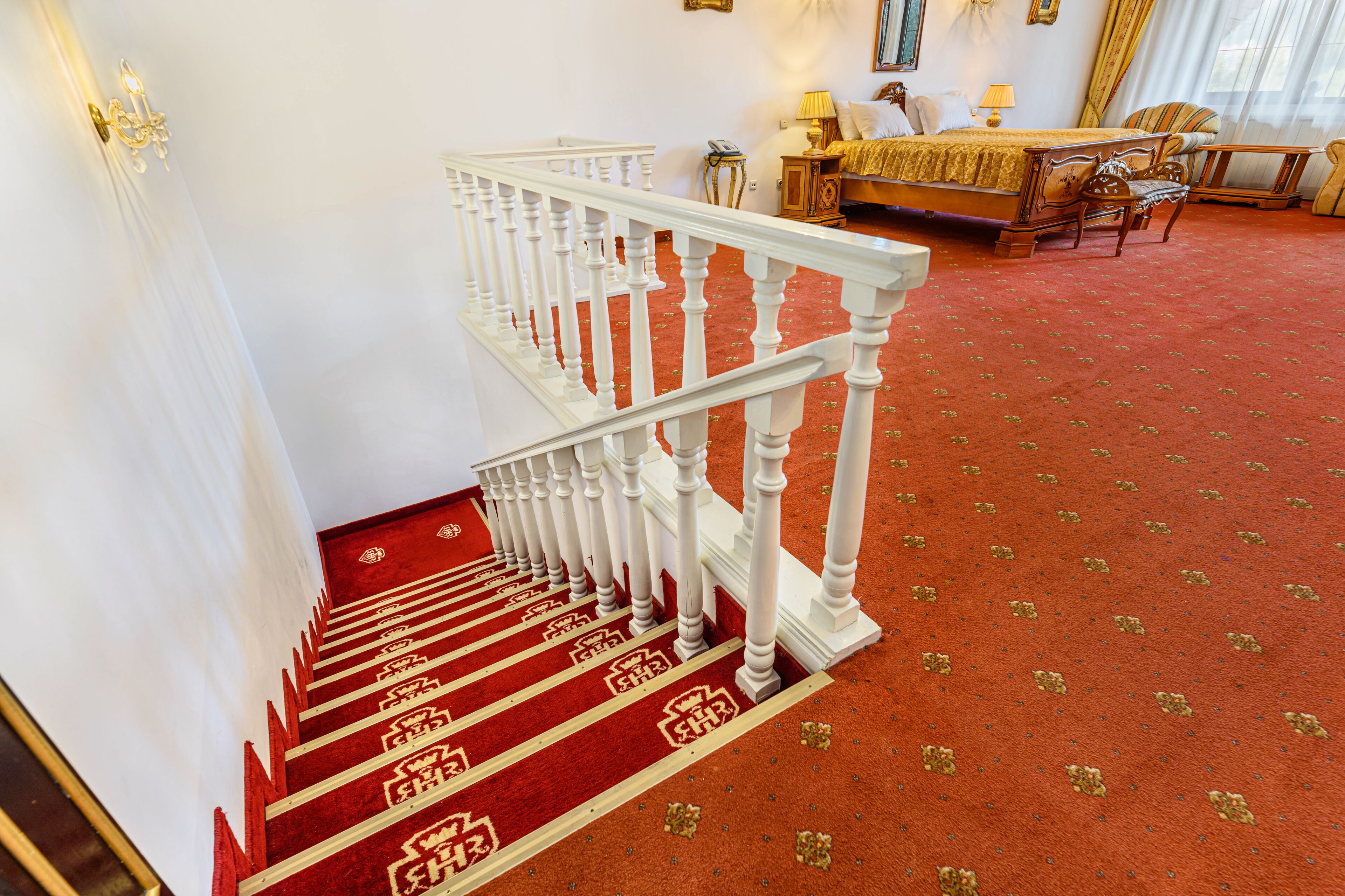 Een statige trap met een rood tapijt en een witte balustrade die leidt naar een slaapkamer met een bed, nachtkastje en lamp.