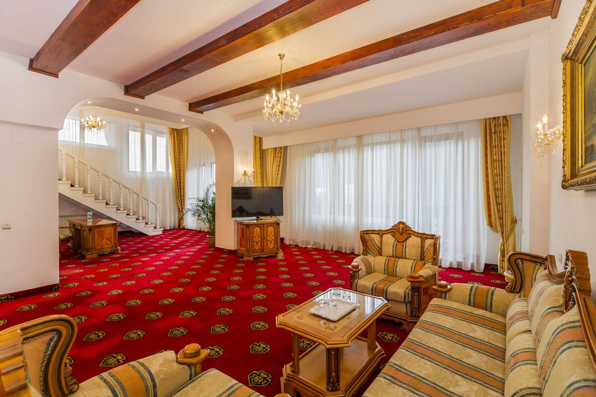 Amplia sala de estar con alfombra roja, muebles de madera, una araña y una escalera que sube a otro nivel.