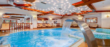 Una amplia piscina interior con caída de agua, vigas de madera y varias zonas de estar.