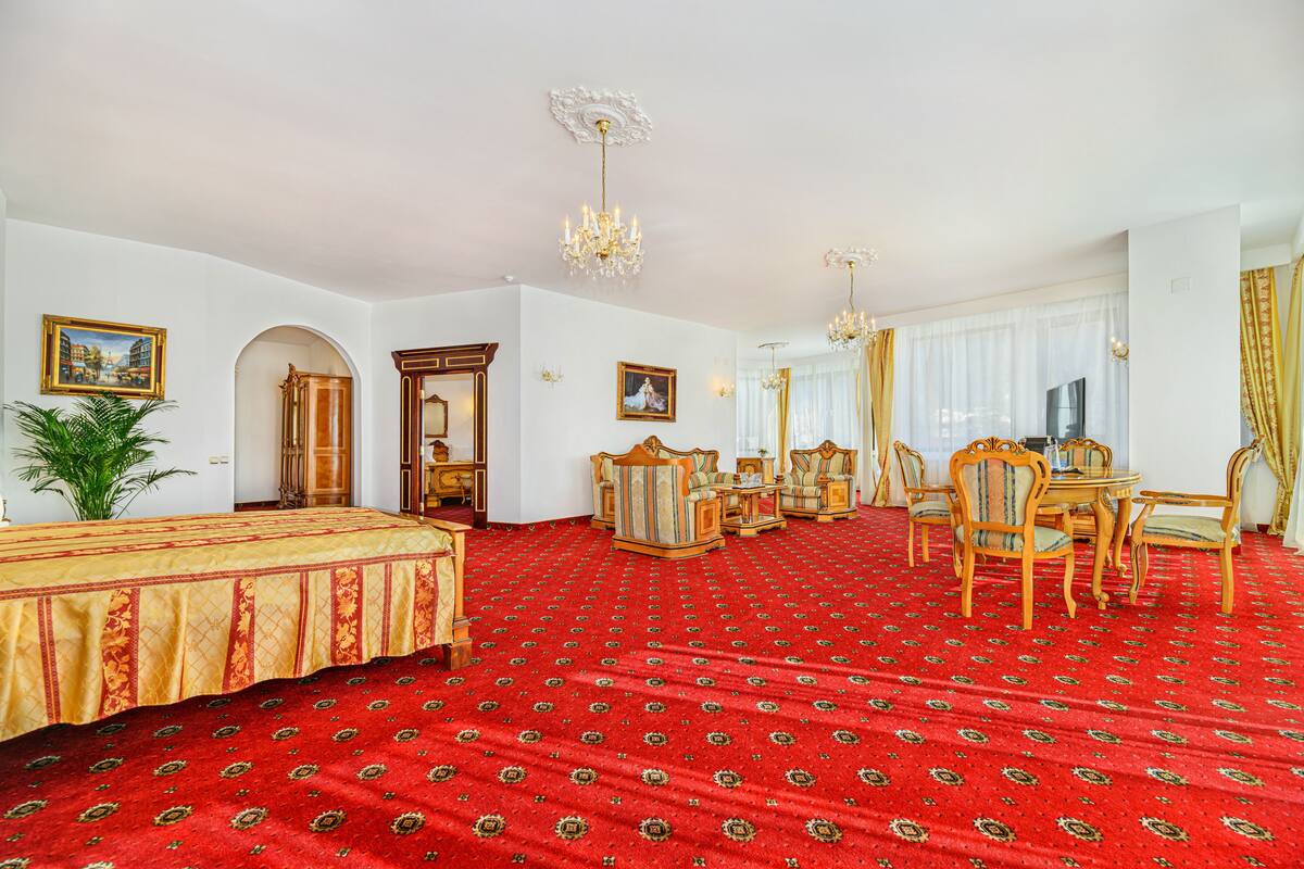 Habitación de hotel con alfombra roja, cama, mesa de comedor con sillas, televisión y un espejo grande.