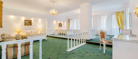 A hotel room with a green patterned carpet, white furniture, a balcony with a white railing, and a chandelier.