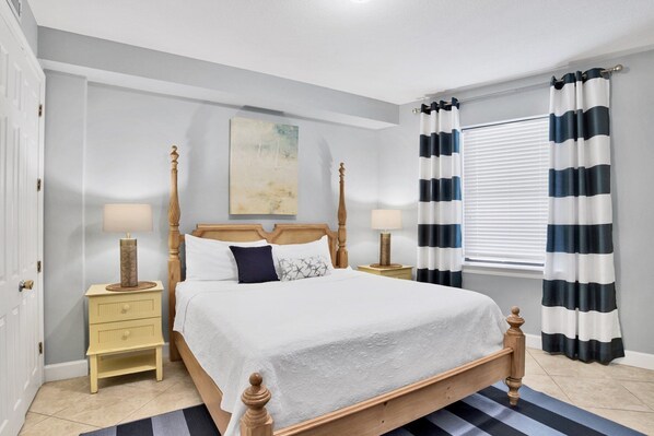 A bedroom with a four-poster bed, two bedside tables, a window with striped curtains, and a painting on the wall.