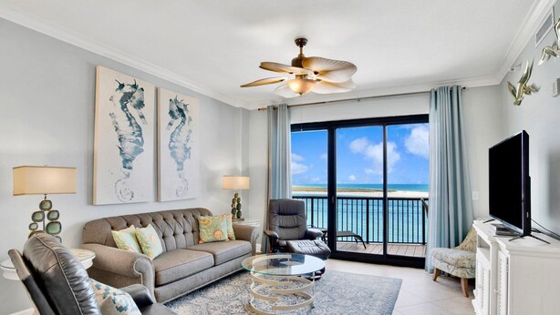 Ein Wohnzimmer mit einer Couch, Sesseln, einem Couchtisch und Kunstwerken an der Wand. Es gibt einen Fernseher und einen Deckenventilator. Das Zimmer verfügt über ein großes Fenster mit Blick aufs Meer.