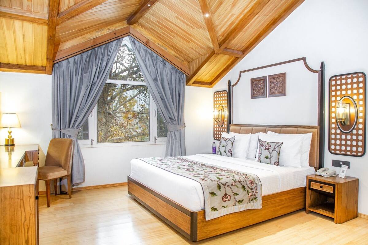 Quarto com cama grande, teto de madeira, escrivaninha com cadeira e janela com cortinas.