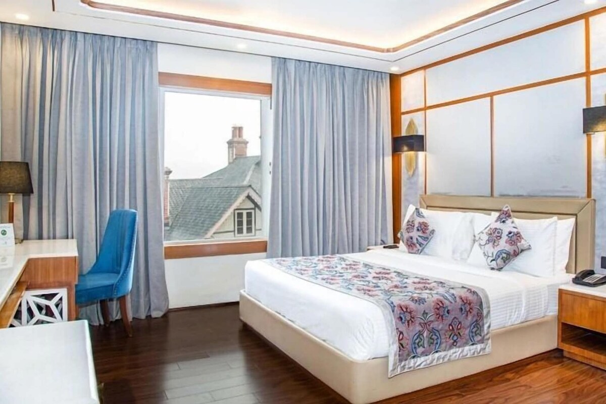 A hotel room with a bed, a desk, a chair, and a view of a house through the window.