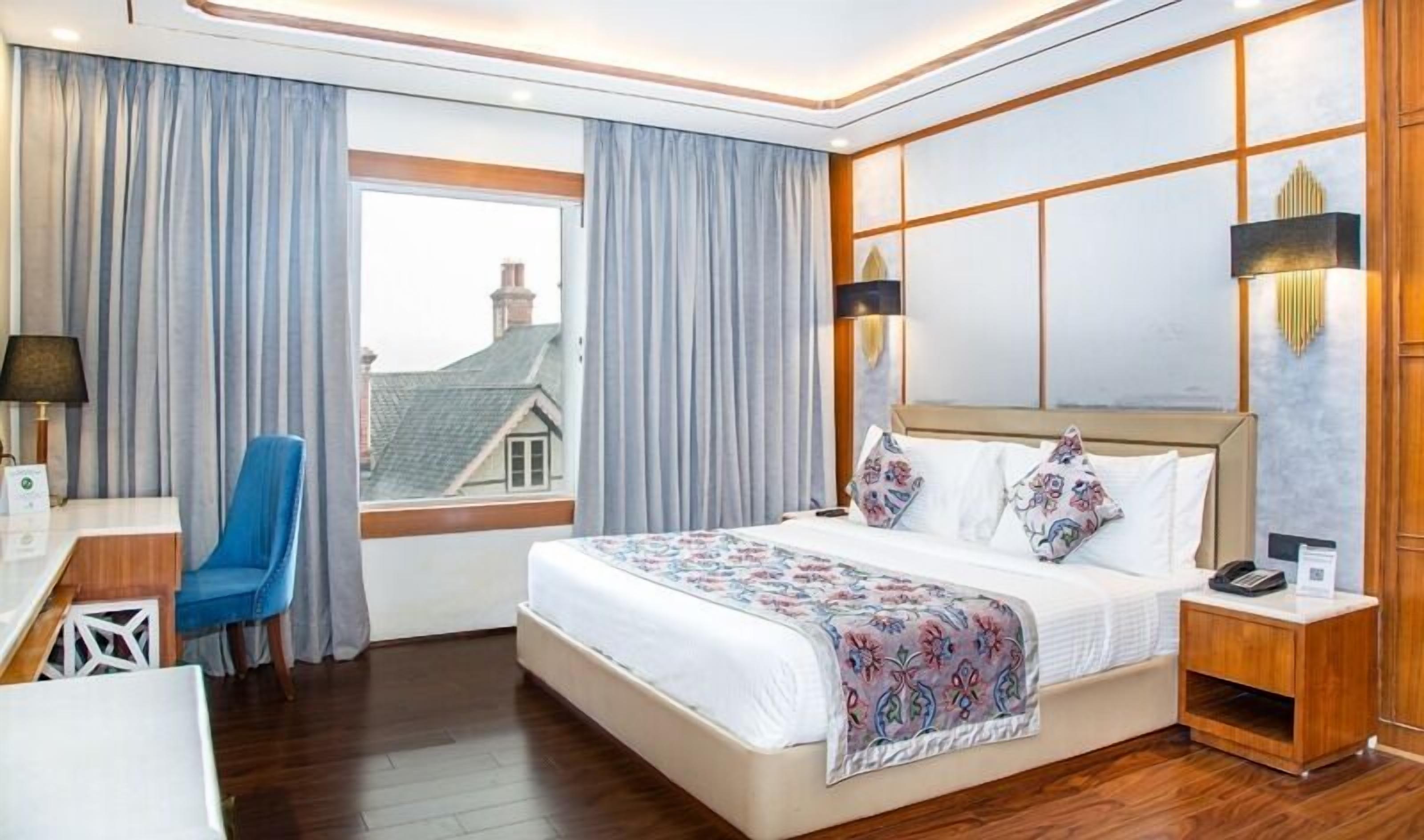 A hotel room with a large bed, a desk, a chair, and a view of a house through the window.
