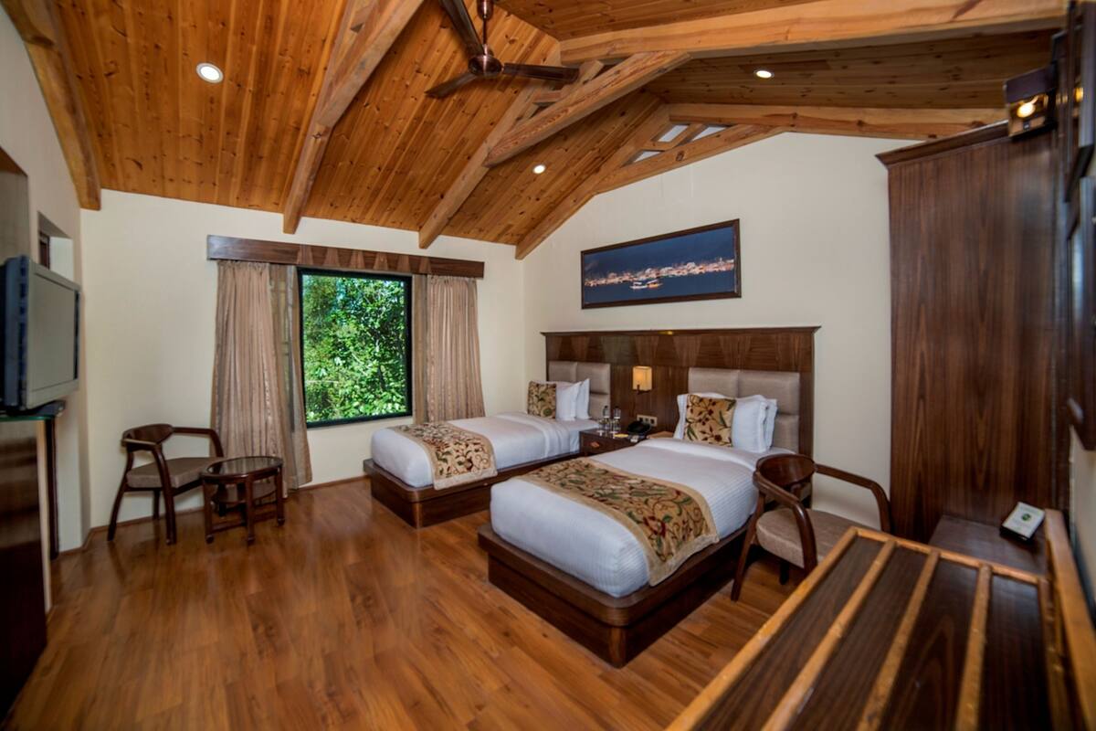 Quarto de hotel com duas camas, piso de madeira, ventoinha no teto, uma janela grande com cortinas e televisão montada na parede.