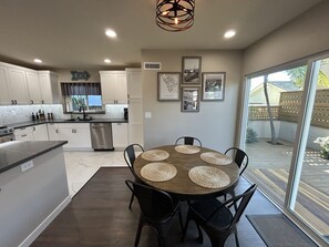 Uma cozinha moderna com armários brancos, forno embutido e máquina de lavar louça. Há uma área de jantar com mesa redonda e cadeiras pretas, e uma grande janela com vista para o quintal.